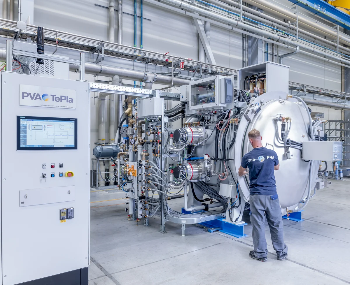 A technician operates a large vacuum sintering furnace system in a bright factory setting, with control cabinets and complex piping visible around the equipment.
