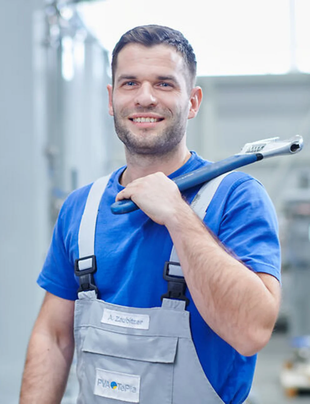 Smiling worker in overalls holds a large wrench while standing in a bright industrial facility