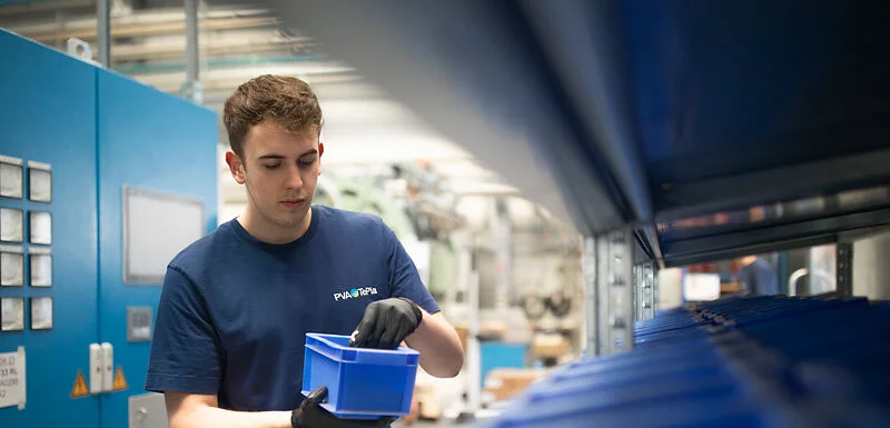 A person wearing work gloves and a blue shirt stands in an industrial workspace, inspecting a blue plastic container. Rows of similar containers and machinery surround them, creating a clean, organized production environment.