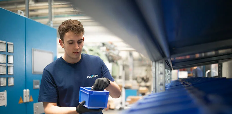 A person wearing work gloves and a blue shirt stands in an industrial workspace, inspecting a blue plastic container. Rows of similar containers and machinery surround them, creating a clean, organized production environment.