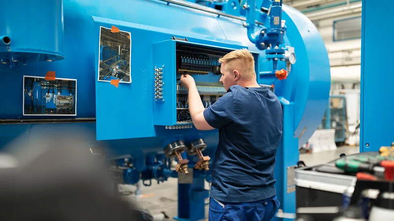 Worker in a factory setting adjusts components inside a large blue vacuum furnace.