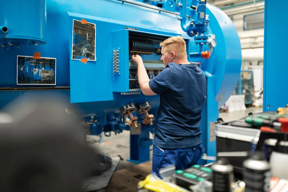 Worker in a factory setting adjusts components inside a large blue vacuum furnace.