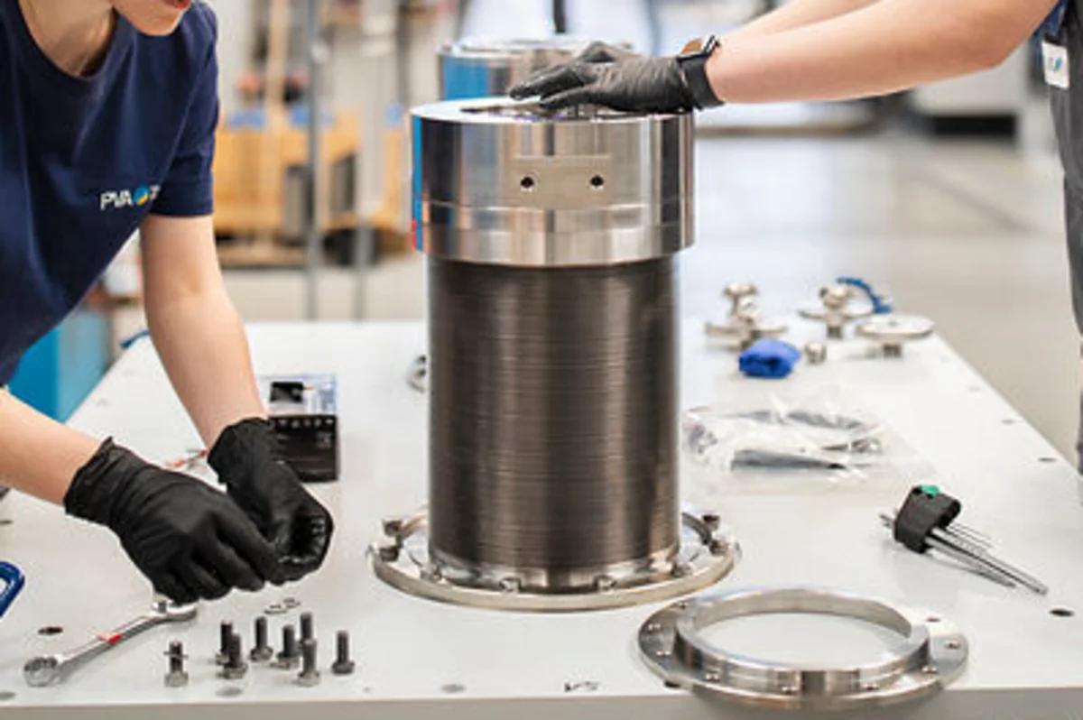 Two workers assemble metal components on a table in a bright industrial workshop.