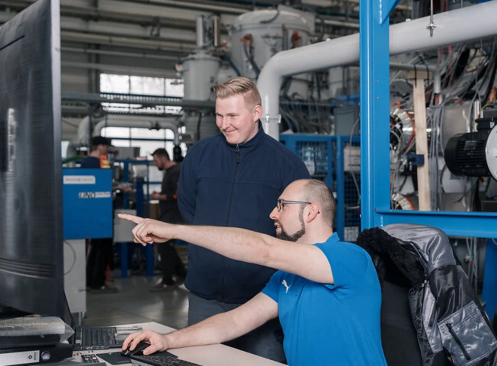 Two workers in an industrial facility review information on a monitor while discussing machine details