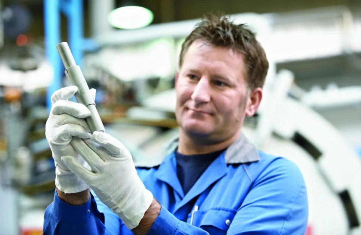 Worker in a blue uniform carefully inspects a cylindrical metal part in an industrial setting.
