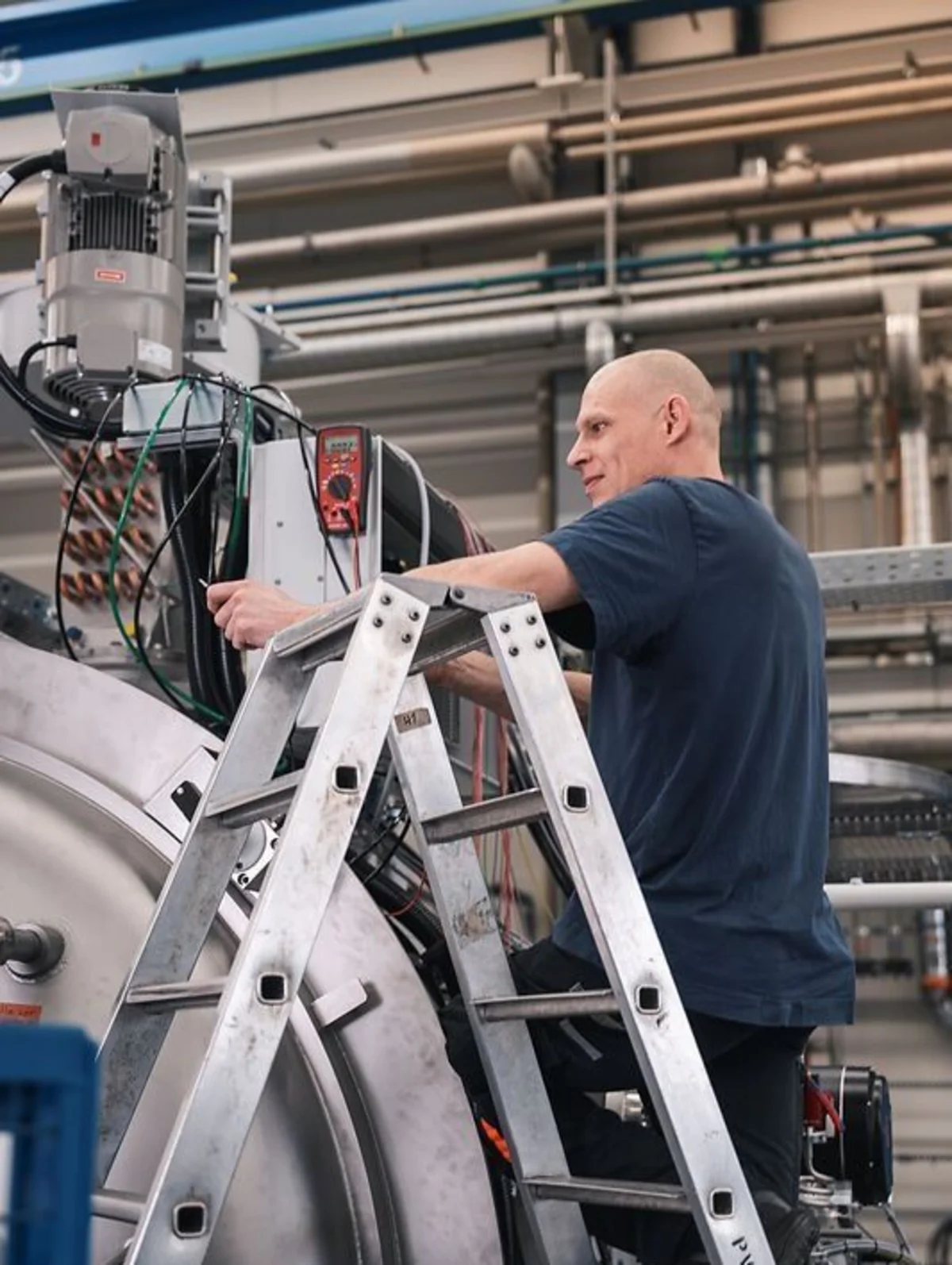 Worker on a ladder adjusts components on a large industrial machine inside a spacious production hall.