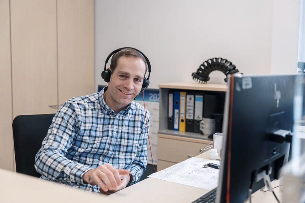 Person with headset sits at an office desk, smiling while working on a computer in a modern workspace.