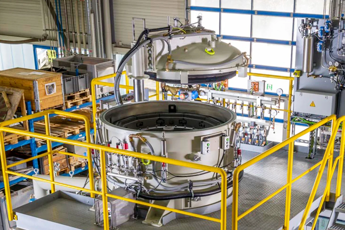 Large vacuum furnace with open dome lid mounted on a yellow support frame in a production facility.