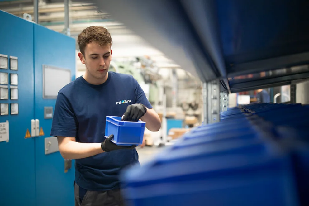 A person wearing work gloves and a blue shirt stands in an industrial workspace, inspecting a blue plastic container. Rows of similar containers and machinery surround them, creating a clean, organized production environment.
