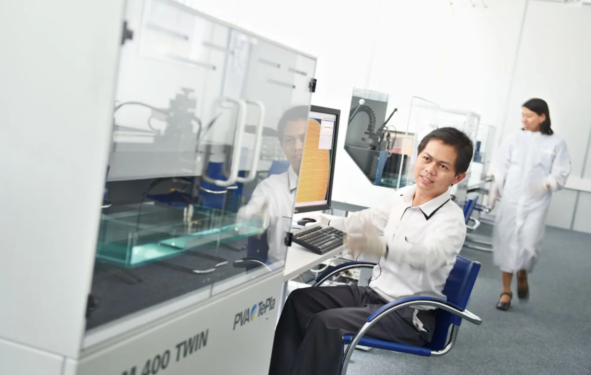 Lab worker operates scanning acoustic microscopy process in a clean, bright laboratory while another person walks in the background.