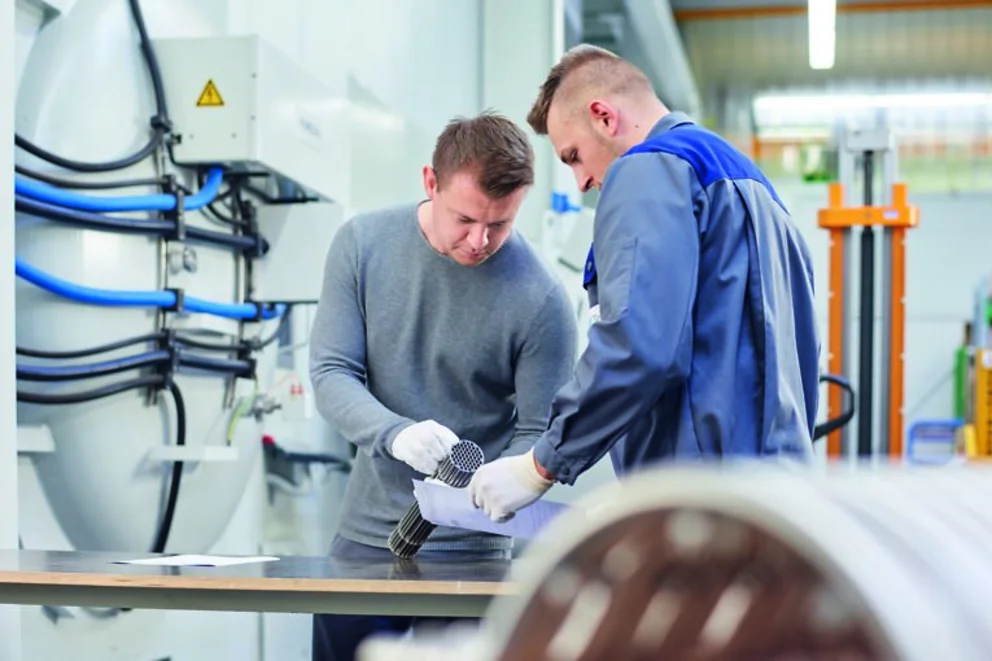 Two workers examine metal parts on a table, discussing details in a bright industrial workshop.