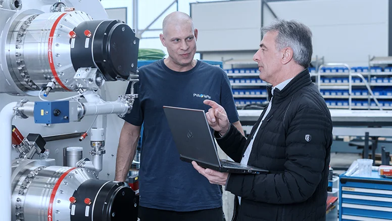 Two workers discuss details of a large industrial machine while reviewing information on a laptop.