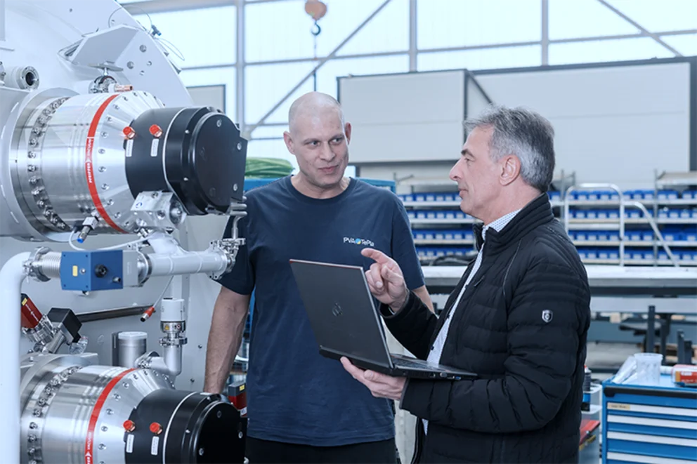 Two workers discuss details of a large industrial machine while reviewing information on a laptop.