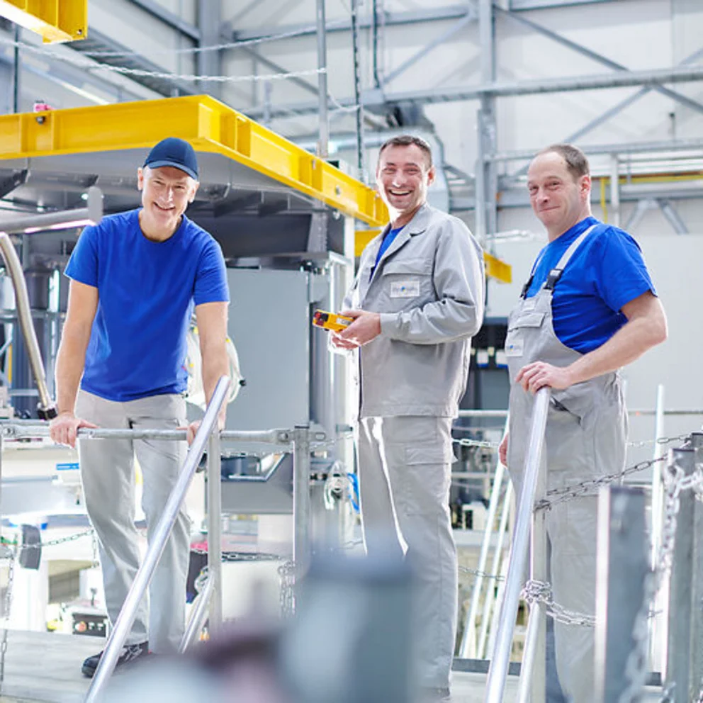 Three workers stand on a platform in a bright industrial hall, inspecting and working on a crystal growing system.