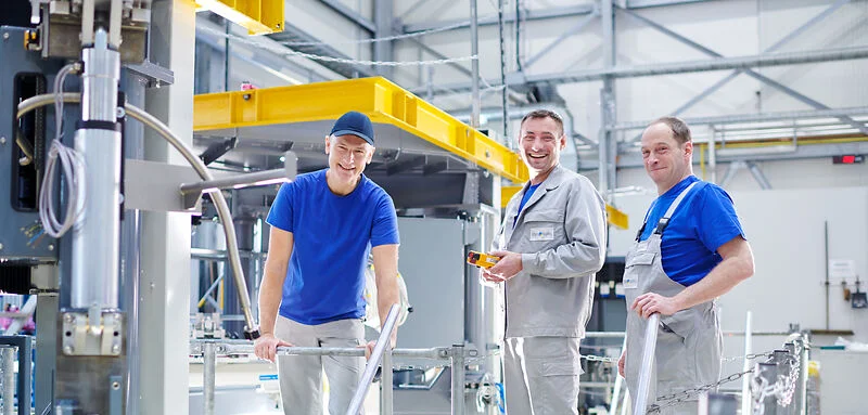 Three workers stand on a platform in a bright industrial hall, inspecting and working on a crystal growing system.