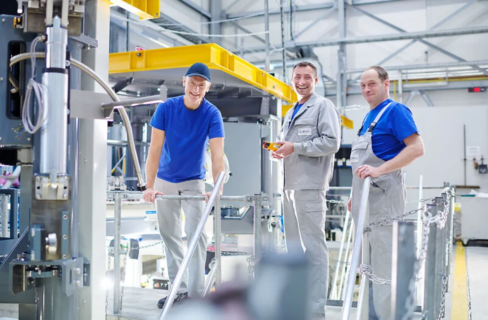 Three workers stand on a platform in a bright industrial hall, inspecting and working on a crystal growing system.