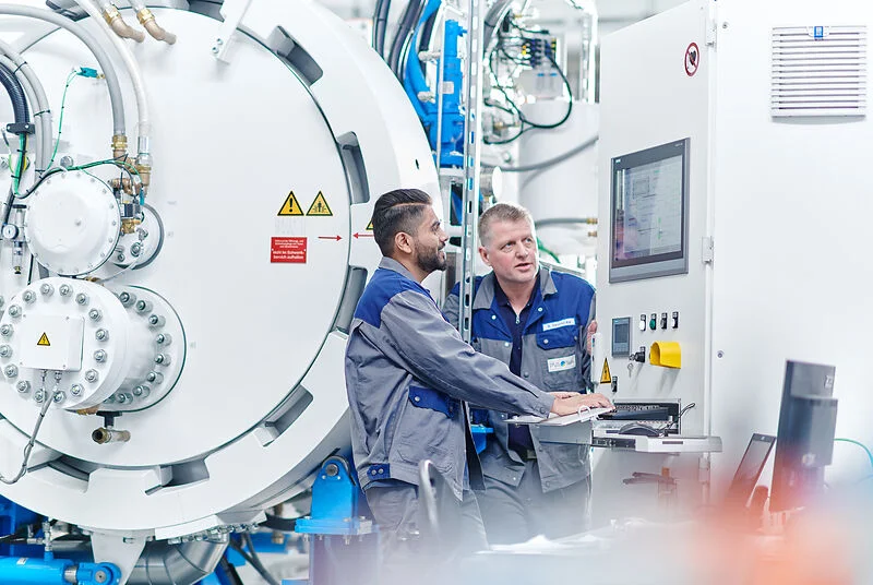 Two workers operate a control panel beside a large industrial furnace, discussing machine settings.